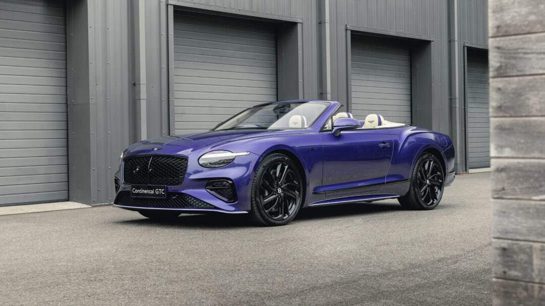 A dynamic 4K wallpaper features a striking dark purple Bentley Continental GT Speed Convertible with its top down, parked on asphalt in front of a sleek, grey industrial building with multiple garage doors. Its glossy, deep purple finish and luxurious cream leather interior, visible with the roof down, create a powerful and sophisticated contrast against the muted, urban backdrop.