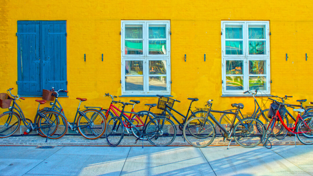 A lively 4K wallpaper featuring a row of diverse bicycles parked neatly against a strikingly vibrant yellow building in Copenhagen, Denmark. The bold contrast of the building's sunny yellow hue against deep blue shutters and the cast shadows on the light pavement creates a picturesque urban scene.