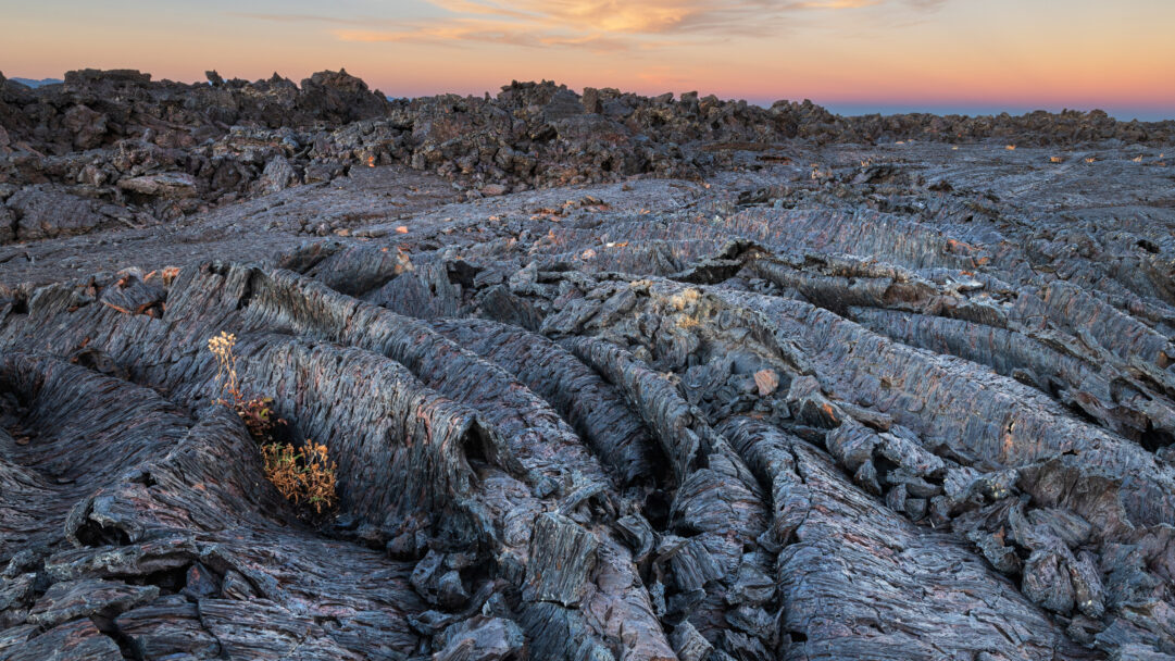 A captivating 4K wallpaper presents the Blue Dragon Lava Flow at Craters of the Moon National Monument, stretching across a vast and rugged landscape. This ancient, dark blue-gray pahoehoe lava, characterized by its flowing, rope-like patterns, is bathed in the soft, warm light of a sunset sky shifting from peach to deep lavender.