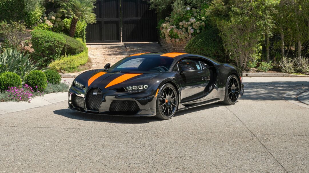 A dynamic 4K wallpaper showcasing a Bugatti Chiron Super Sport 300+ in black with bold orange racing stripes, parked on a sunlit asphalt driveway in front of a grand black gate framed by vibrant green foliage. The car's glossy finish reflects the bright daylight, emphasizing its powerful lines and the vivid contrast of the orange accents against the deep black body.