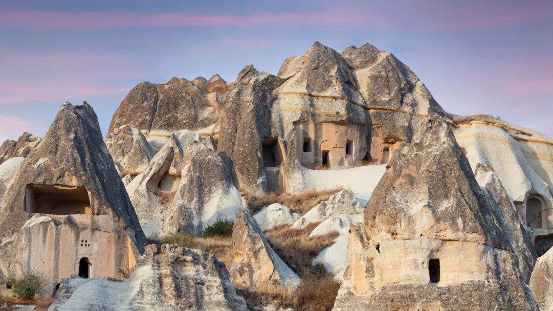 A magical 4K wallpaper of Cappadocia's unique landscape, featuring numerous fairy chimneys with ancient cave dwellings carved into their sides. Soft, diffused sunlight illuminates the intricate rock textures and the mysterious entrances of the cave homes, all set against a tranquil, pastel-streaked dawn sky.