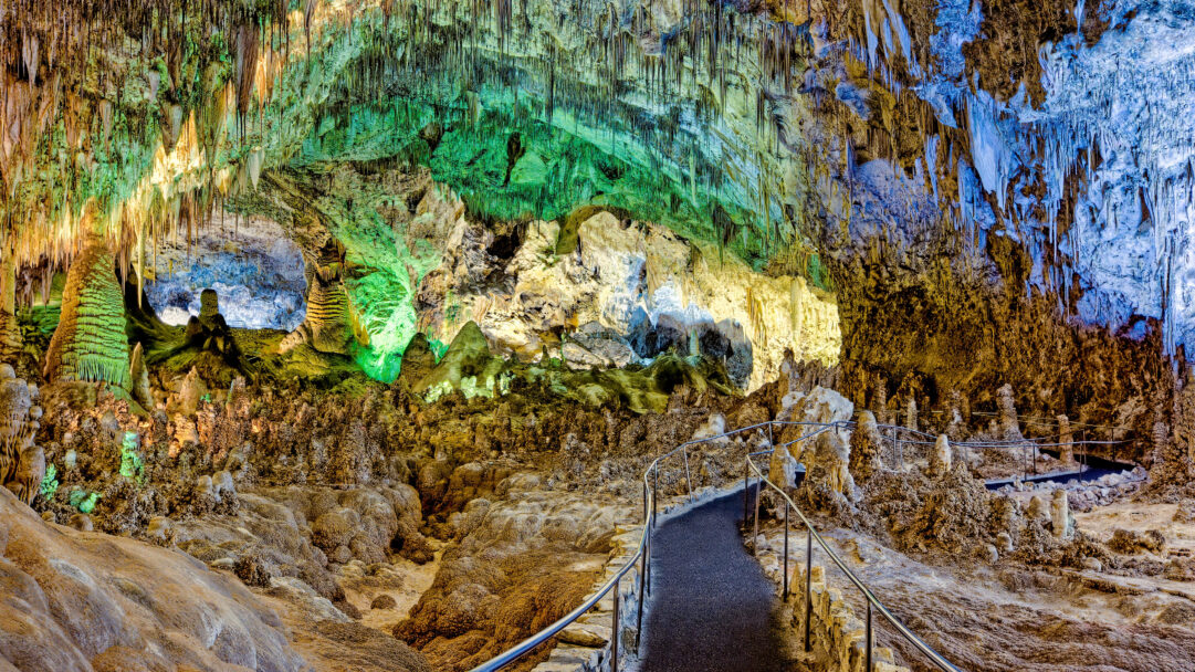 A breathtaking 4K wallpaper showcases the vast Big Room of Carlsbad Caverns, with an illuminated walkway winding through an immense chamber of geological formations. Vibrant spotlights cast ethereal greens, yellows, and blues across countless stalactites, stalagmites, and flowstones, creating an otherworldly and awe-inspiring subterranean landscape.