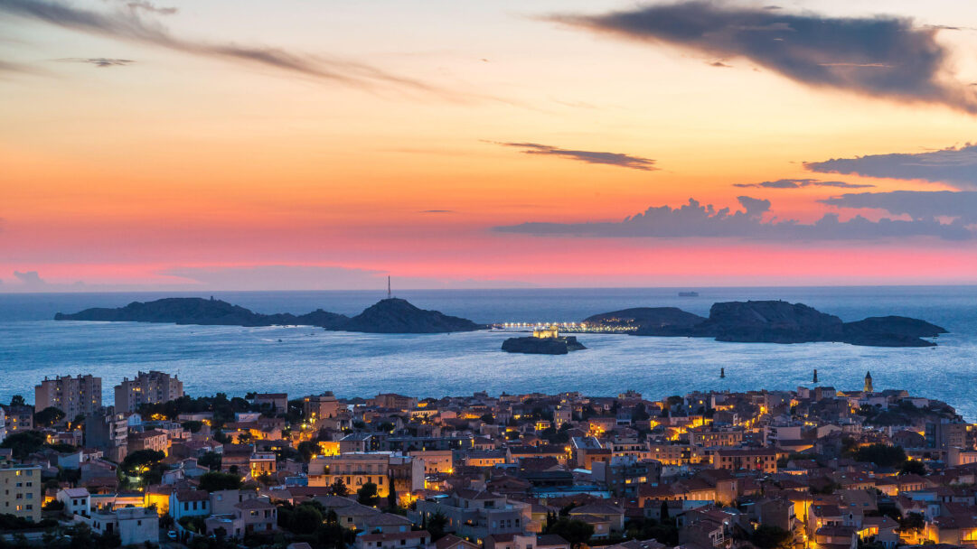 A breathtaking 4K wallpaper depicts the historic Château D'if within the expansive Marseille Port and city, bathed in the tranquil light of dusk. The vibrant sunset sky transitions from warm oranges to deep pinks, illuminating the sparkling city lights and the dark silhouette of the fortress against the shimmering sea.
