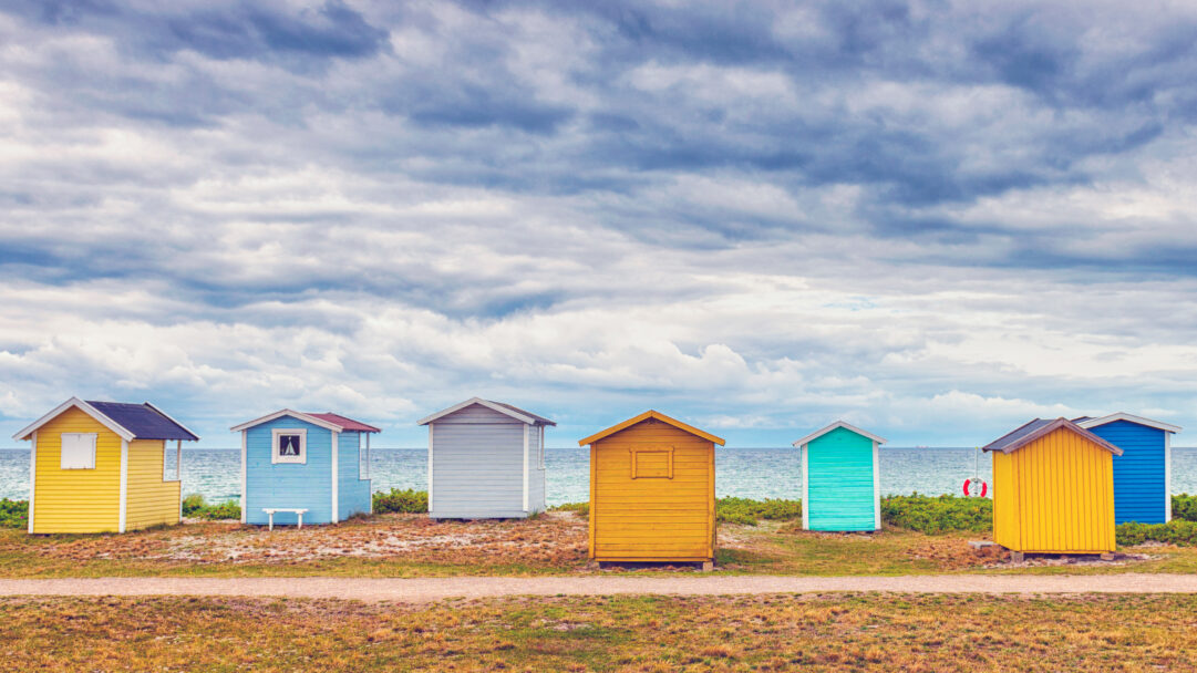 A charming 4K wallpaper of a row of colorful bathing huts stretching along a sandy Skåne beach path under a vast, cloudy sky. Each hut, painted in distinct shades of yellow, blue, orange, and turquoise, adds a cheerful pop of color against the serene, muted tones of the distant sea and dramatic overcast sky.