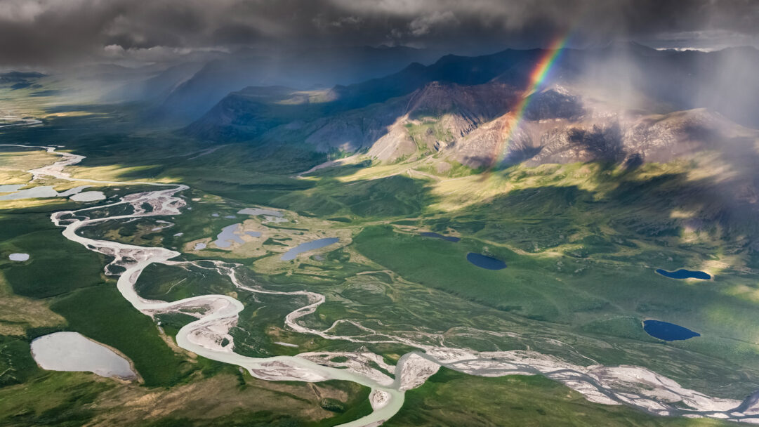 A majestic 4K wallpaper showcasing an aerial view of the wide, braided confluence of Easter Creek and Killik River flowing through a vast green valley, interspersed with small lakes and framed by distant mountains. A brilliant rainbow arcs vibrantly from dramatic dark clouds, illuminating sun-dappled peaks and casting striking light and shadow patterns across the expansive, rugged landscape.