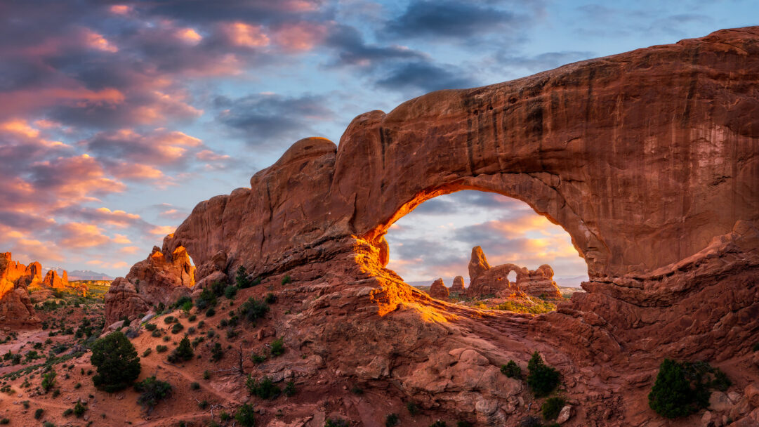 A breathtaking 4K wallpaper showcases the North Window Turret Arch formation in Arches National Park, Utah, bathed in the warm evening light. The vibrant sunset hues intensely highlight the striking red sandstone and the rugged desert landscape, contrasting with a dramatic sky of glowing orange, pink, and deep blue clouds.