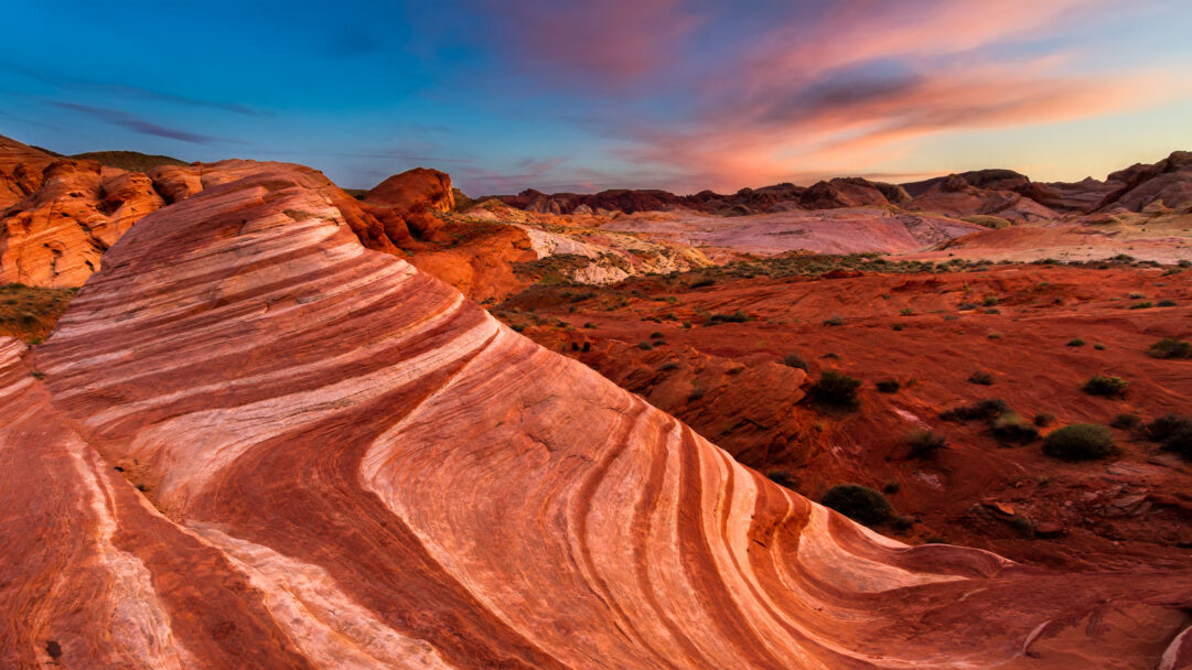An awe-inspiring 4K wallpaper presents the iconic Fire Wave rock formation at sunset within Valley of Fire State Park. Vivid stripes of fiery red and white undulate across the foreground, mirrored by a dramatic sky painted with hues of deep blue, soft pink, and warm orange.