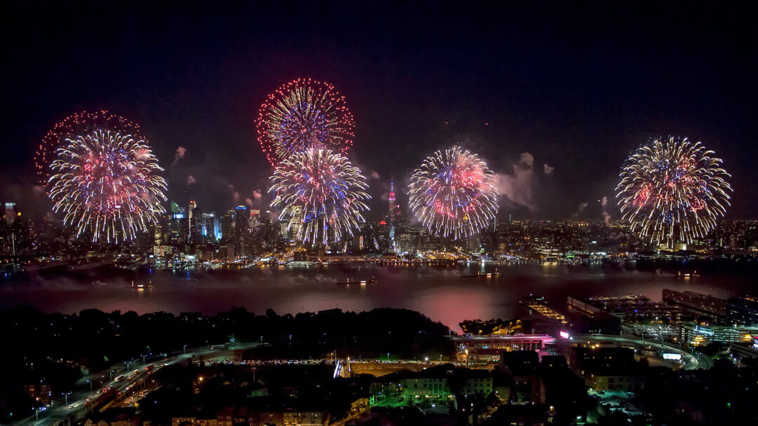 A spectacular 4K wallpaper capturing Fourth of July fireworks exploding brilliantly over the Hudson River, illuminating the iconic New York City skyline. Vibrant bursts of red, white, and blue light up the night sky, casting colorful reflections on the water and highlighting the city's festive glow.