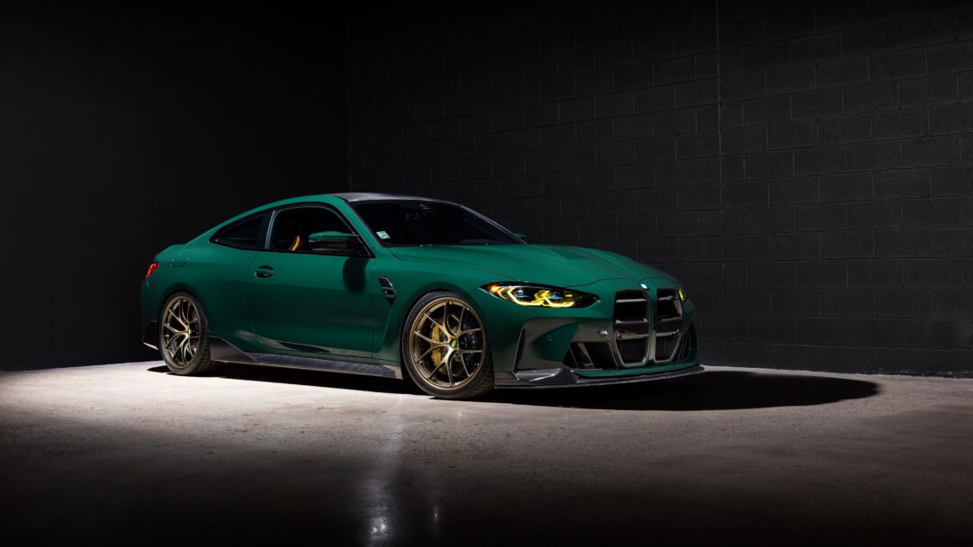 An exhilarating 4K wallpaper showcases a sleek, green BMW M4 Coupe with striking gold wheels, dynamically parked against a dark, industrial brick wall background. The car's vibrant green finish and glowing yellow headlights dramatically contrast with the shadowed concrete floor and dark surroundings, creating an intense, high-performance mood.