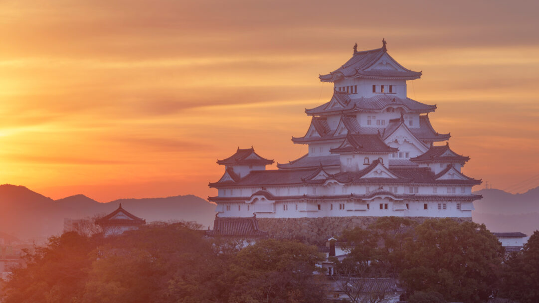 A breathtaking 4K wallpaper featuring Himeji Castle, a historic white fortress, situated atop a hill in Himeji, Hyogo Prefecture, Japan. The castle is dramatically silhouetted against a brilliant sunset sky, painted with hues of warm orange and gold, evoking a sense of ancient splendor.