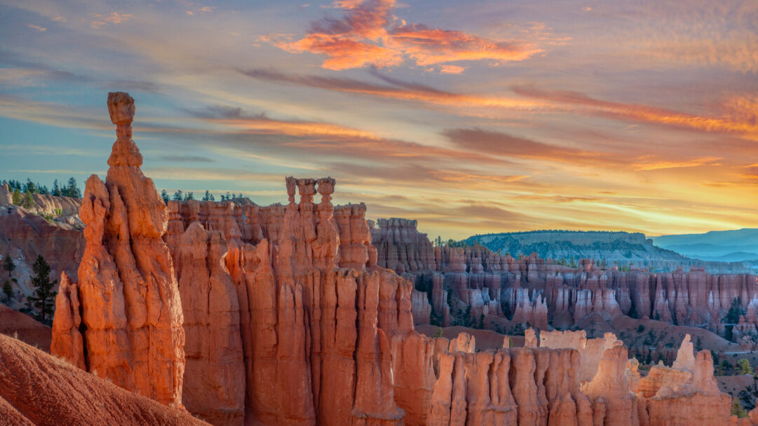 A breathtaking 4K wallpaper captures the majestic hoodoos of Bryce Canyon at Sunset Point, presenting a vast landscape of eroded rock formations under a vivid sky. The golden hour light bathes the unique orange and pink rock pillars, creating a dramatic contrast against the fiery, layered clouds streaking across the vast horizon.