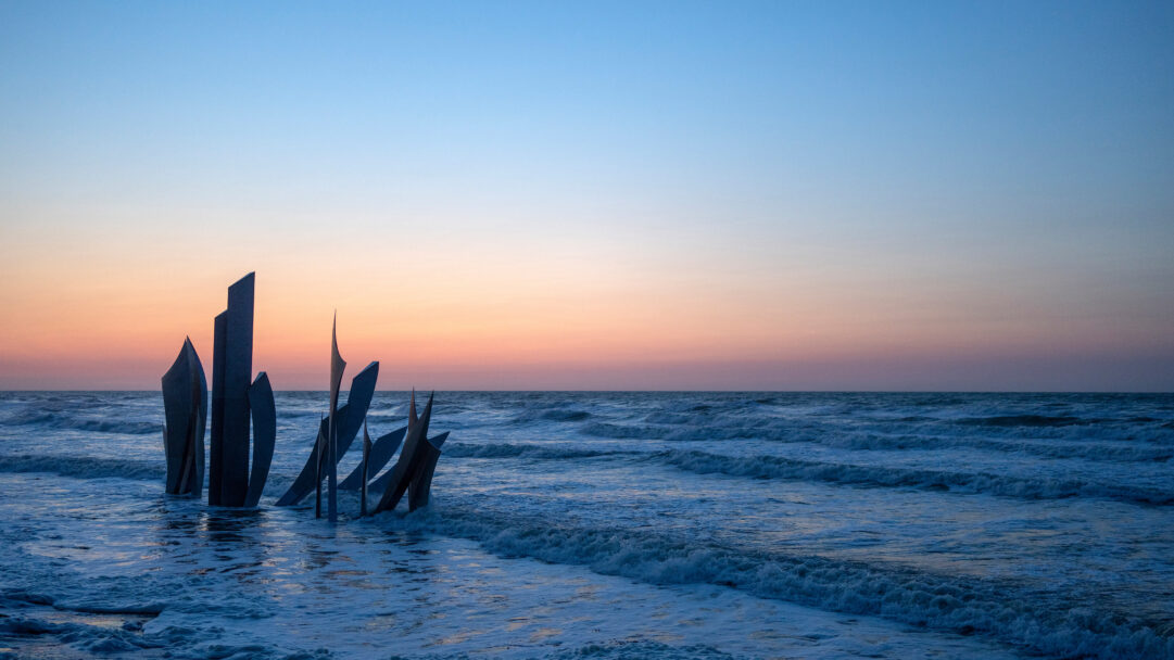 An impactful 4K wallpaper captures the powerful Les Braves Monument standing amidst the crashing waves of Omaha Beach at sunset. The monument's dark, angular forms are silhouetted against a sky painted with soft hues of orange and pink, beautifully reflected in the deep blue, frothy waves.