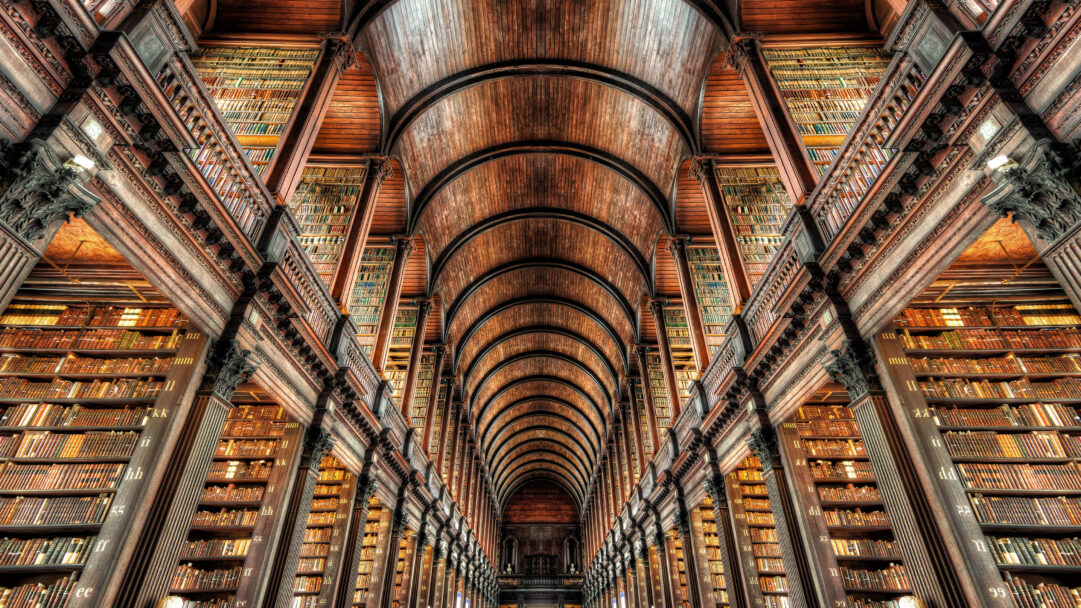 A captivating 4K wallpaper displays the majestic Long Room of the Library of Trinity College Dublin, Ireland, filled with rows upon rows of ancient books stretching towards a high arched wooden ceiling. The symmetrical perspective emphasizes the room's immense scale and historic grandeur, with warm light illuminating the richly textured wooden architecture and countless leather-bound volumes.
