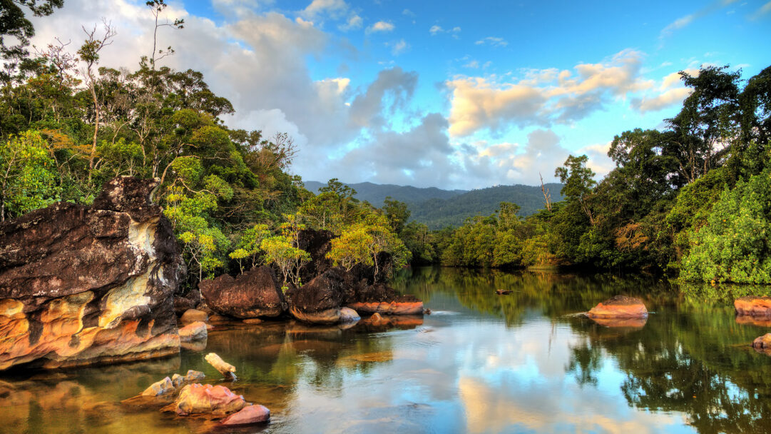 A breathtaking 4K wallpaper showcasing the stunning river, forest, and rock landscape of Masoala National Park in Madagascar. Warm sunlight illuminates the vibrant green foliage and rich reddish-brown rocks, which are mirrored in the calm water under a dramatic blue and cloudy sky.