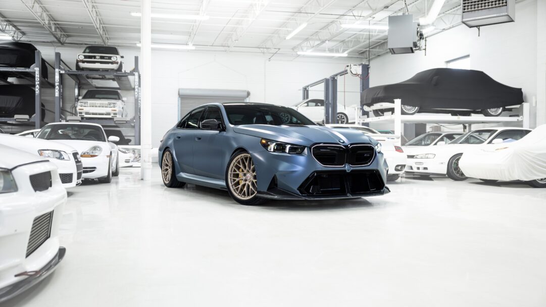 An exquisite 4K wallpaper showcasing a matte blue BMW M5 sedan with striking gold wheels, prominently parked in a pristine, brightly lit garage filled with other luxury vehicles. The crisp lighting and polished white floor enhance the car's bold design, emphasizing its luxurious and powerful presence amidst the sophisticated automotive collection.