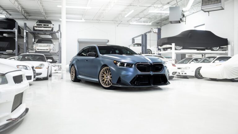 An exquisite 4K wallpaper showcasing a matte blue BMW M5 sedan with striking gold wheels, prominently parked in a pristine, brightly lit garage filled with other luxury vehicles. The crisp lighting and polished white floor enhance the car's bold design, emphasizing its luxurious and powerful presence amidst the sophisticated automotive collection.