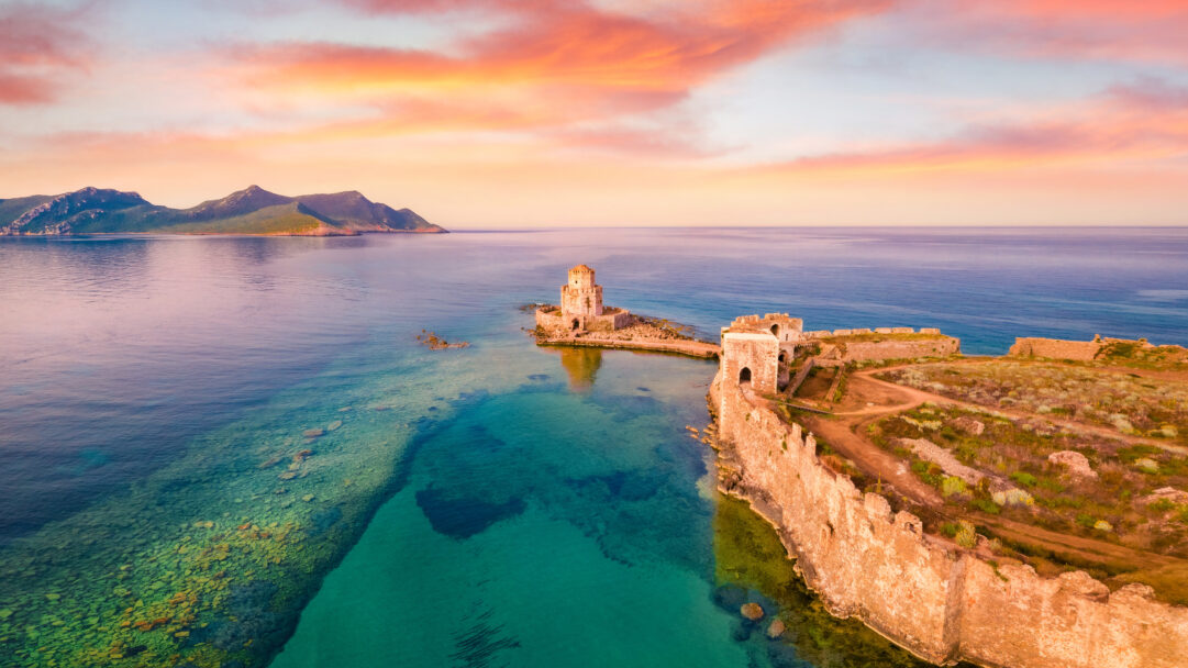 A captivating 4K wallpaper showcasing the ancient Methoni Castle ruins in Messenia, Greece, perched on a coastal peninsula with its octagonal Bourtzi fortress on an adjacent islet. The vivid hues of the sunset paint the sky in oranges and pinks, brilliantly reflecting upon the impossibly clear, multi-toned turquoise waters surrounding the historic stone fortifications.