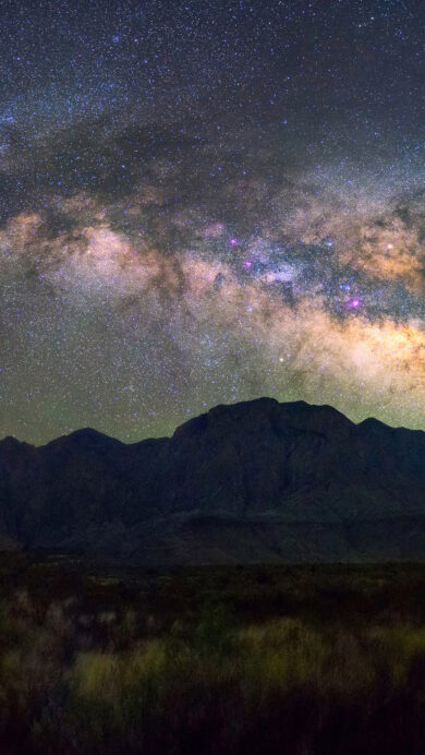 An awe-inspiring 4K wallpaper capturing the brilliant arc of the Milky Way Galaxy stretching across a vast, star-filled night sky above the silhouetted peaks of Big Bend National Park mountains. Vibrant cosmic dust lanes of purple, gold, and green illuminate the heavens, contrasting dramatically with the dark, rugged landscape below, creating a mood of profound nocturnal majesty.