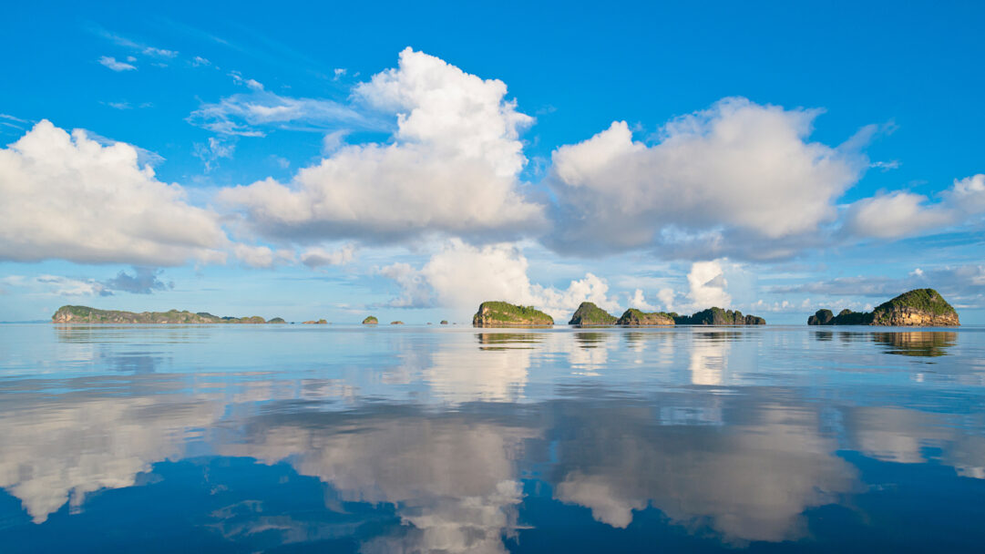A breathtaking 4K wallpaper presents the majestic Misool Raja Ampat Islands of Indonesia scattered across a vast, calm ocean under a brilliant blue sky. The tranquil water perfectly mirrors the fluffy white clouds and blue expanse above, creating a serene, immersive double landscape defined by peaceful reflections.