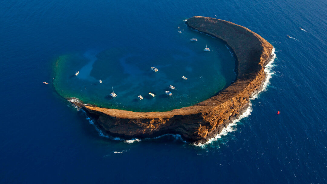 A breathtaking 4K wallpaper captures an aerial view of Molokini Crater, a crescent-shaped islet off the coast of Maui, Hawaii, surrounded by the vast blue Pacific Ocean. Inside its sheltered lagoon, numerous boats dot the clear turquoise waters, inviting exploration of the vibrant coral reefs illuminated by golden sunlight.