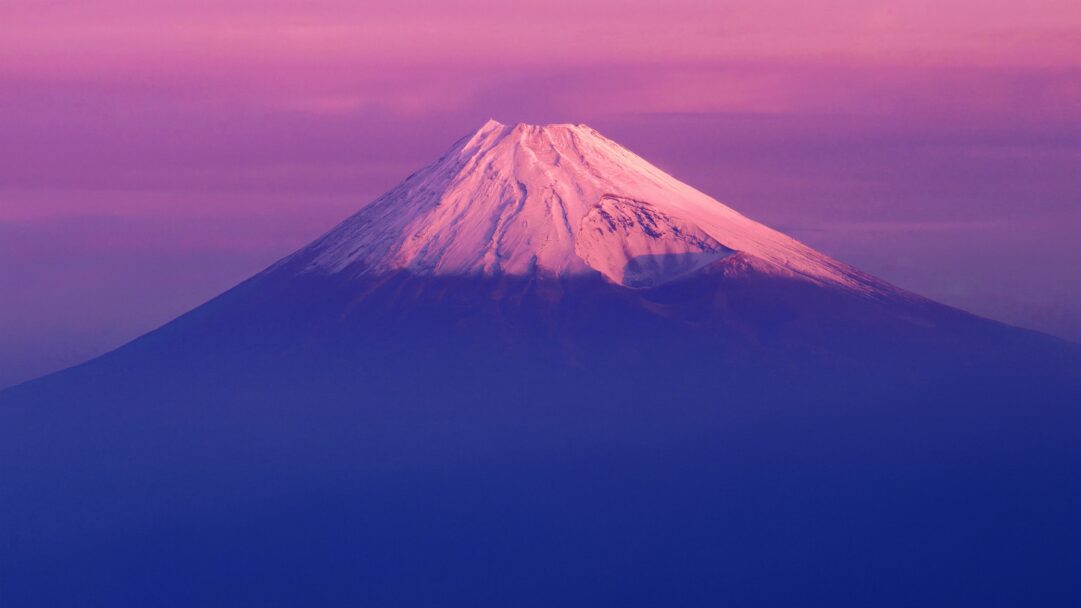 A breathtaking 4K wallpaper depicts the iconic OS X Lion Mount Fuji, its snow-capped peak rising majestically against a vibrant pink and purple gradient sky. The mountain's crisp white and pink-tinged snow catches a soft light, beautifully contrasted by the deep violet and rose hues of the expansive atmosphere, evoking a sense of calm grandeur.