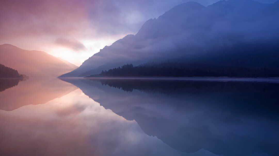 A breathtaking 4K wallpaper captures a tranquil lake nestled among majestic, mist-shrouded mountains, a scene often inspiring the serene desktop environments of OS X. The striking interplay of soft pink and purple hues from the sunrise transitions to deep blues on the right, all perfectly mirrored in the glassy lake surface, evoking a profound sense of peace.