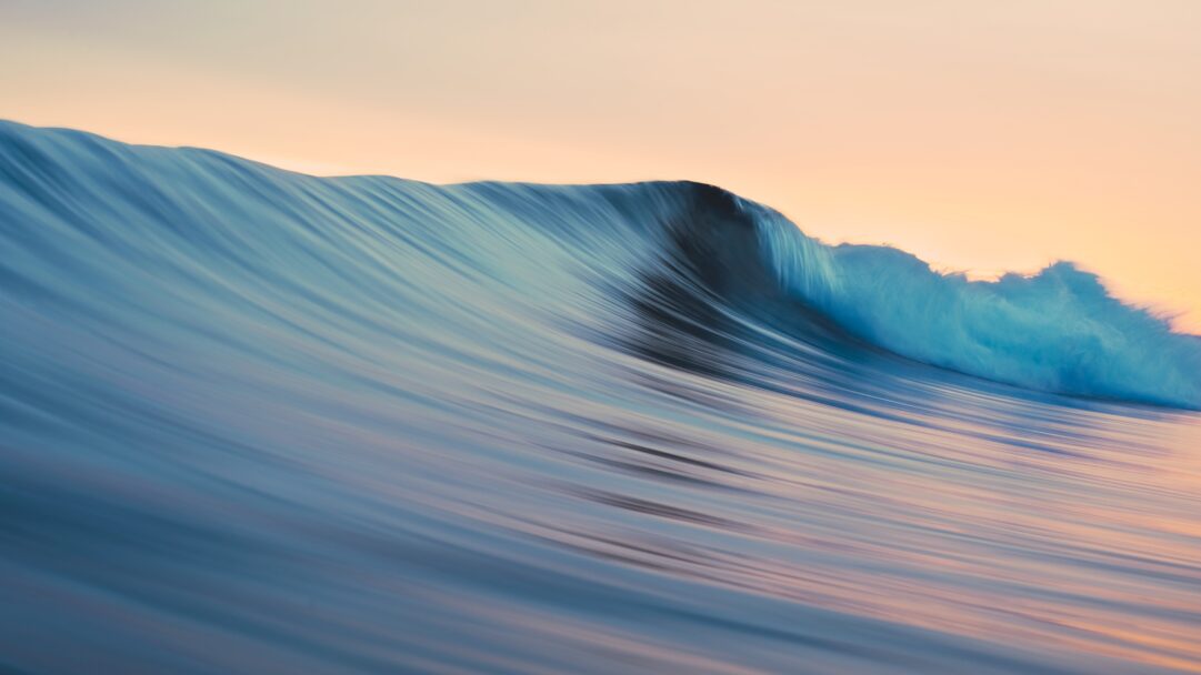 A mesmerizing 4K wallpaper of the iconic OS X Mavericks blue wave, captured in motion as it crests across the frame under a warm, diffused sky. Its translucent turquoise crest glows brightly against the soft orange and pink hues of the sky, with the water's surface displaying a serene, blurred texture from its flowing movement.