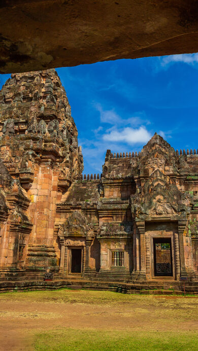 A majestic 4K wallpaper offers a stone-framed vista of the Prasat Phanom Rung Temple ruins, revealing its impressive sandstone structure under an expansive blue sky. Sunlight illuminates the temple's intricate carvings and warm reddish-brown hues, contrasting with the vibrant blue sky and the dark, moss-covered foreground stone.