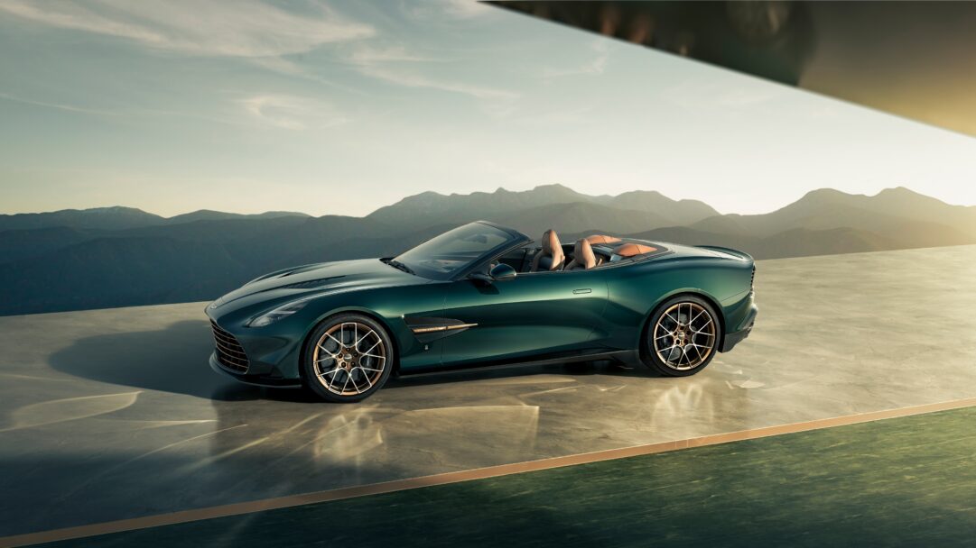 An exquisite 4K wallpaper capturing a dark green Q by Aston Martin Vanquish Volante convertible parked on a sophisticated platform, set against a majestic mountain range at sunset. The car's deep metallic green finish beautifully reflects the warm, golden light of the declining sun, creating a luxurious and serene mood across the dramatic landscape.