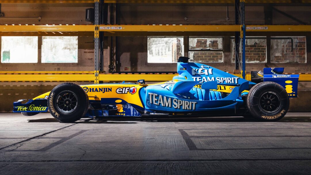 A captivating 4K wallpaper showcasing the iconic Renault R25 F1 car, prominently displaying its blue and yellow livery, positioned inside a utilitarian warehouse. Dramatic lighting emphasizes the car's sleek aerodynamic form and vibrant sponsor decals, creating a striking visual against the muted concrete floor and industrial yellow shelving.