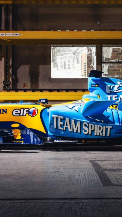 A captivating 4K wallpaper showcasing the iconic Renault R25 F1 car, prominently displaying its blue and yellow livery, positioned inside a utilitarian warehouse. Dramatic lighting emphasizes the car's sleek aerodynamic form and vibrant sponsor decals, creating a striking visual against the muted concrete floor and industrial yellow shelving.