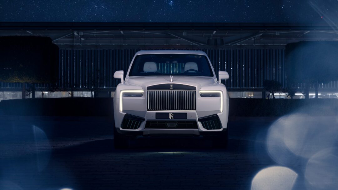 A celestial 4K wallpaper showcasing a pristine white Rolls-Royce Cullinan SUV parked head-on at night beneath a brilliant, star-studded cosmos. Its illuminated grille and prominent daytime running lights pierce the serene darkness, complemented by subtle reflections on the paved surface and the elegant architecture in the background.