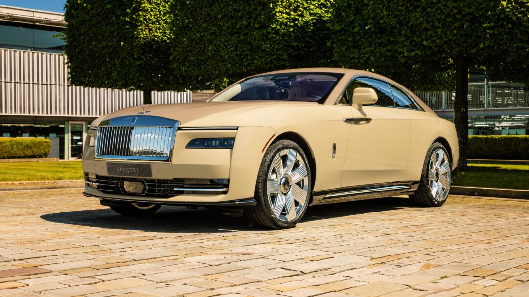 An opulent 4K wallpaper showcasing the Rolls-Royce Spectre Bailey 2025, a luxurious light beige electric coupe, parked elegantly on a stone-paved drive in front of a modern building and lush green foliage. Its highly polished chrome grille and intricately designed wheels gleam brilliantly under bright sunlight, reflecting the clear sky and emphasizing the vehicle's sophisticated craftsmanship and serene presence.