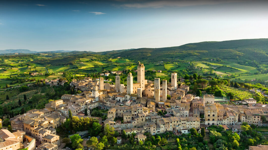 An exquisite 4K wallpaper showcasing the ancient town of San Gimignano with its historic towers rising prominently from a hilltop, nestled within the sprawling, verdant landscape of Siena, Tuscany. Golden afternoon light bathes the medieval stone buildings and their tall structures, creating a stunning contrast with the vibrant green vineyards and rolling hills under a serene blue sky.