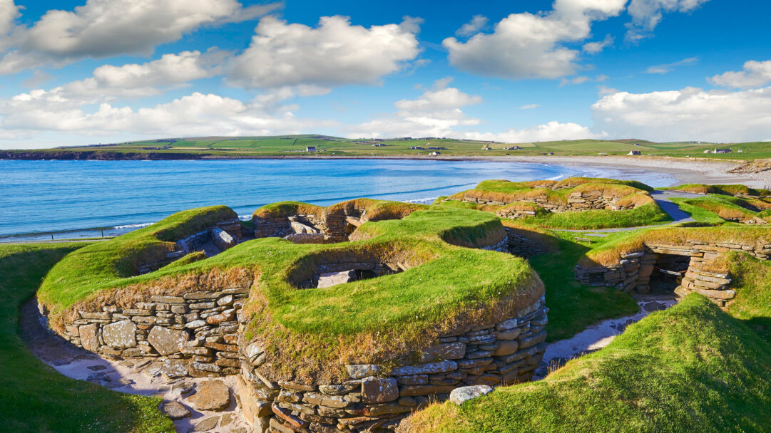 An expansive 4K wallpaper showcasing the Skara Brae Neolithic Settlement nestled on a vibrant green coastal landscape under a bright blue, cloudy sky. The remarkable well-preserved stone houses, partly earth-sheltered and covered in lush grass, contrast beautifully with the calm turquoise sea and distant rolling hills.