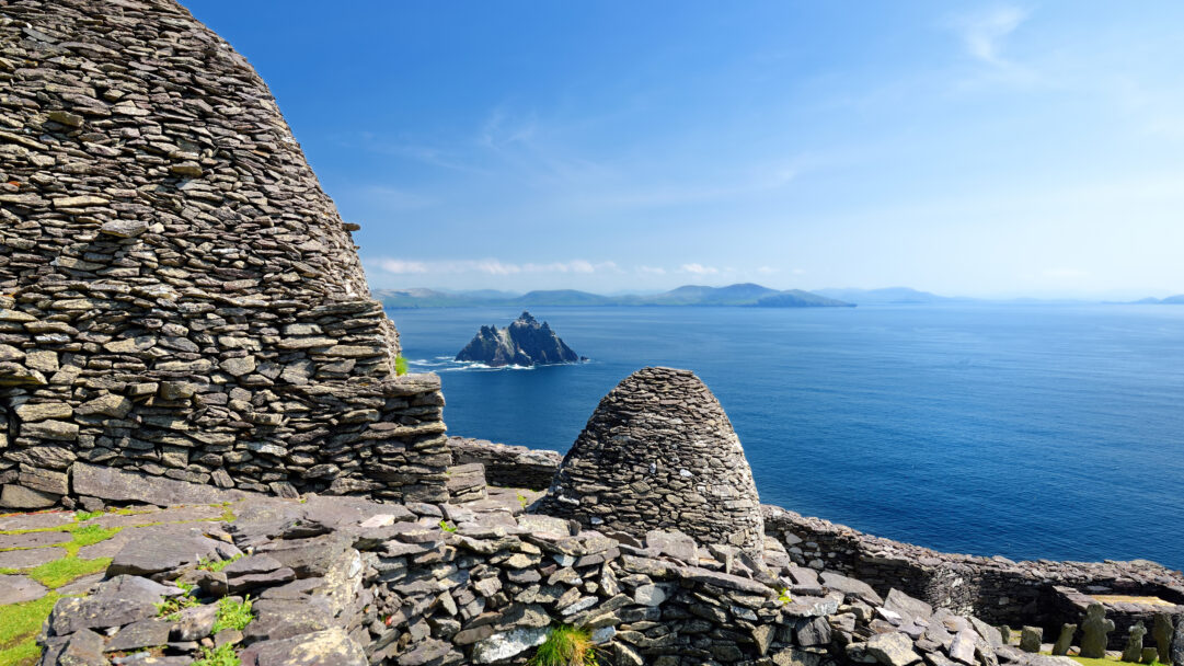 An ancient 4K wallpaper showcases the rugged Skellig Michael monastery ruins, dramatically overlooking the expansive, deep blue Atlantic Ocean. The striking dry-stone structures, illuminated by brilliant sunlight, foreground the distant, mist-shrouded islands and the seemingly endless, shimmering sea.