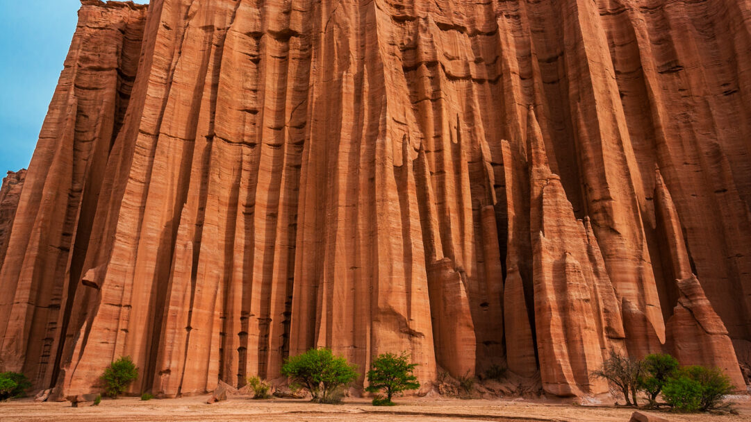 An awe-inspiring 4K wallpaper presents the immense red sandstone cliffs of Talampaya National Park in La Rioja, Argentina, rising dramatically from the desert floor. Their deeply grooved, vertically stratified surfaces showcase rich red-orange hues, emphasizing the ancient, powerful grandeur of this unique geological wonder.