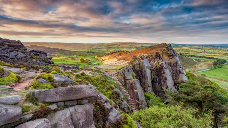 A breathtaking 4K wallpaper showcasing the dramatic rocky landscape of The Roaches in England's Peak District, with a winding path traversing the rugged terrain. Golden light from the setting sun illuminates the textured rock formations and lush green valleys, creating a majestic and serene atmosphere under a sky painted with soft, warm hues.