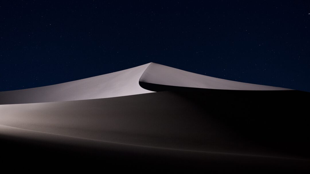 A breathtaking 4K wallpaper presents a colossal sand dune in the Mojave Desert under a profound starry night sky, perfectly suited as a default macOS backdrop. The moonlit crest of the dune glows with stark white precision against the deep indigo expanse, while its dramatically shadowed face plunges into velvety darkness, creating an atmosphere of profound tranquility.