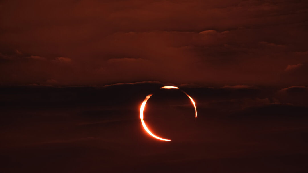 A spectacular 4K wallpaper captures an annular solar eclipse, forming a brilliant 'Ring of Fire' visible against the dark, cloudy sky over Doha, Qatar. The intense, fiery orange ring of the sun glows brightly through parting clouds, casting a dramatic, mystical reddish hue across the darkened celestial expanse.
