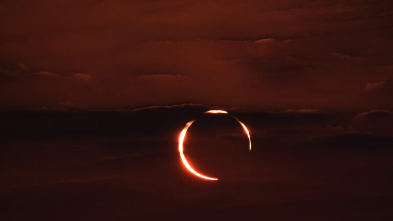 A spectacular 4K wallpaper captures an annular solar eclipse, forming a brilliant 'Ring of Fire' visible against the dark, cloudy sky over Doha, Qatar. The intense, fiery orange ring of the sun glows brightly through parting clouds, casting a dramatic, mystical reddish hue across the darkened celestial expanse.