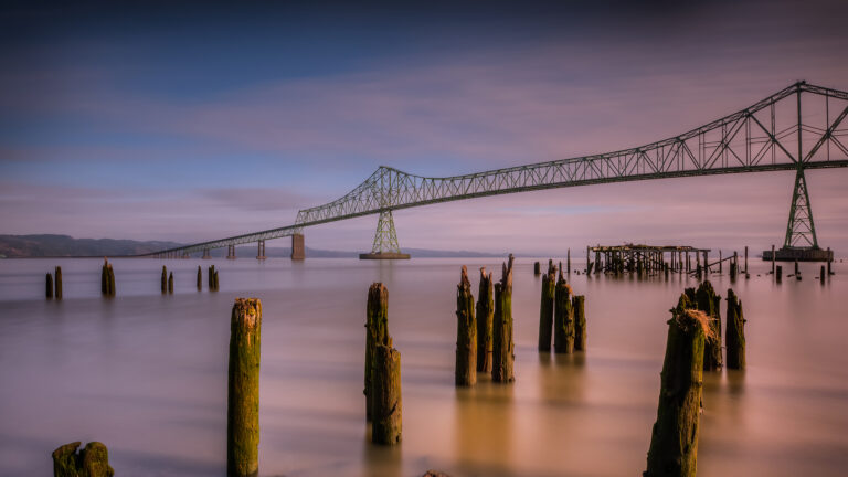 A captivating 4K wallpaper featuring the Astoria-Megler Bridge majestically spanning the Columbia River in Oregon, with weathered wooden pilings emerging from the foreground. The long exposure creates a serene, painterly effect on the water and sky, softly blending twilight hues of muted blue, lavender, and warm amber.