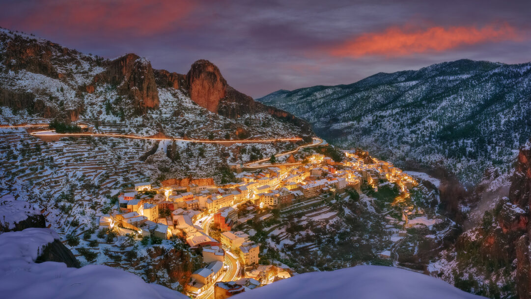 An enchanting 4K wallpaper displaying Aýna Village nestled dramatically into the snowy mountains of Albacete, Spain, under a winter night sky. Warm village lights spill across the snow-dusted rooftops and winding roads, creating a striking contrast with the purpling twilight and dramatic, orange-tinged clouds above.