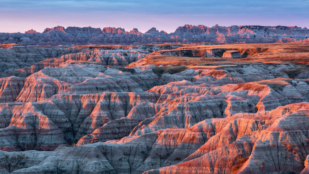 An evocative 4K wallpaper showcases the dramatic, layered formations of Badlands National Park in South Dakota. The setting sun bathes the intricate peaks and valleys in warm golden light, creating a striking contrast with the cool purples and greys of the shadowed areas and the twilight sky.