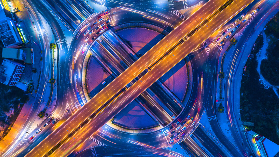 A dazzling 4K wallpaper capturing an aerial long exposure view of a bustling highway interchange and traffic circle in Bangkok, Thailand, at night. The vibrant interplay of orange light trails from vehicles on the elevated highways and blue-purple light trails on the circular roads below creates a dynamic, energetic urban flow.