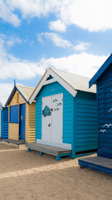 A captivating 4K wallpaper showcasing the famous Bathing Boxes of Brighton Beach, Melbourne, arrayed along a sun-drenched sandy shore. Each box's unique, vivid color scheme stands out sharply under a clear blue sky, casting distinct shadows and embodying a quintessential Australian beach charm.
