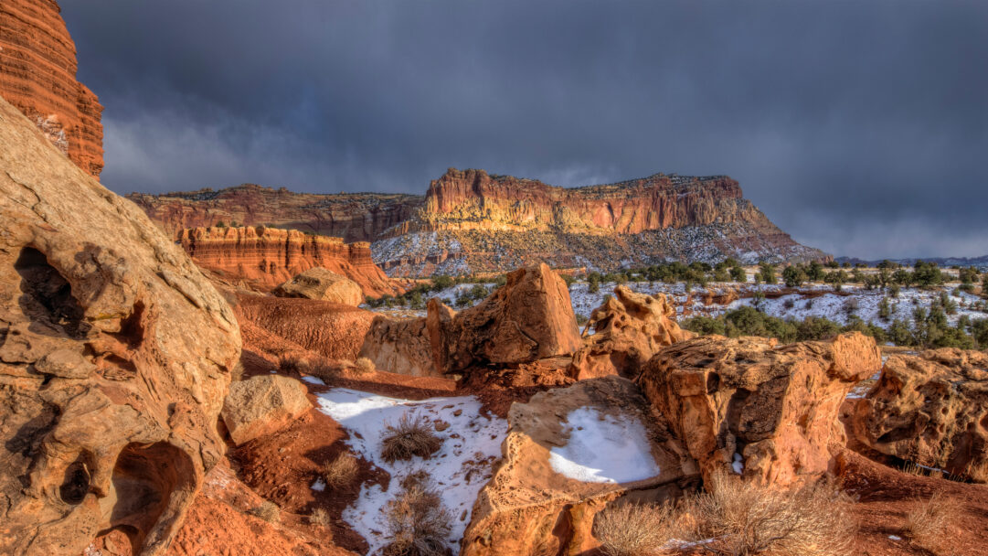 A striking 4K wallpaper showcasing the stark beauty of Capitol Reef National Park, Utah, where towering red rock formations rise from a snow-dusted landscape. Dramatic sunlight breaks through a dark, cloudy sky, brilliantly illuminating distant red cliffs and creating a powerful contrast with the shadowed, snow-dusted foreground.