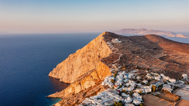 An evocative 4K wallpaper presents an aerial view of the Chora cliffside village on Folegandros Island in the Greek Cyclades. Golden hour sunlight illuminates the rugged, sun-drenched cliffs and the whitewashed buildings, creating a dramatic contrast with the tranquil, deep blue Aegean Sea.