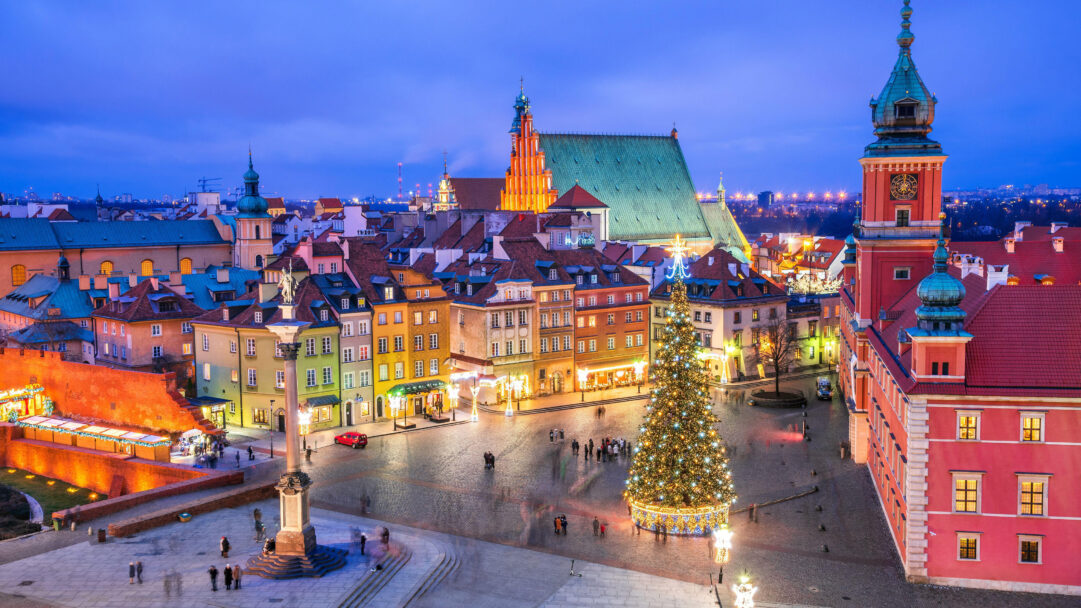 A captivating 4K wallpaper showcases a magnificent, brightly lit Christmas tree centerpiece in the bustling Castle Square of Old Town Warsaw, Poland, surrounded by its charming historic architecture. Golden lights adorn the towering tree and illuminate the vibrant, multi-colored facades of the surrounding buildings, casting a warm, festive glow against the deep twilight blue sky and cobblestone square.