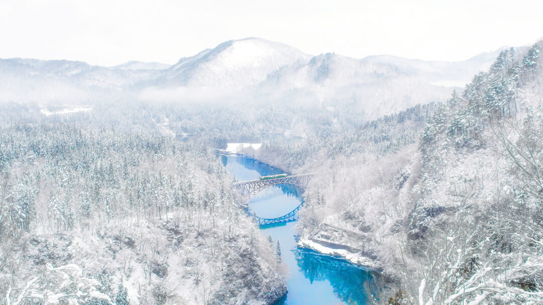 A picturesque 4K wallpaper features the iconic Daiichi Tadami River Bridge in Fukushima, Japan, spanning a bright blue river within a vast, snow-covered winter landscape. A small green and white train is visible crossing the steel bridge, its vibrant colors offering a striking contrast to the surrounding pristine white forests and mountains and the deep turquoise of the river below.
